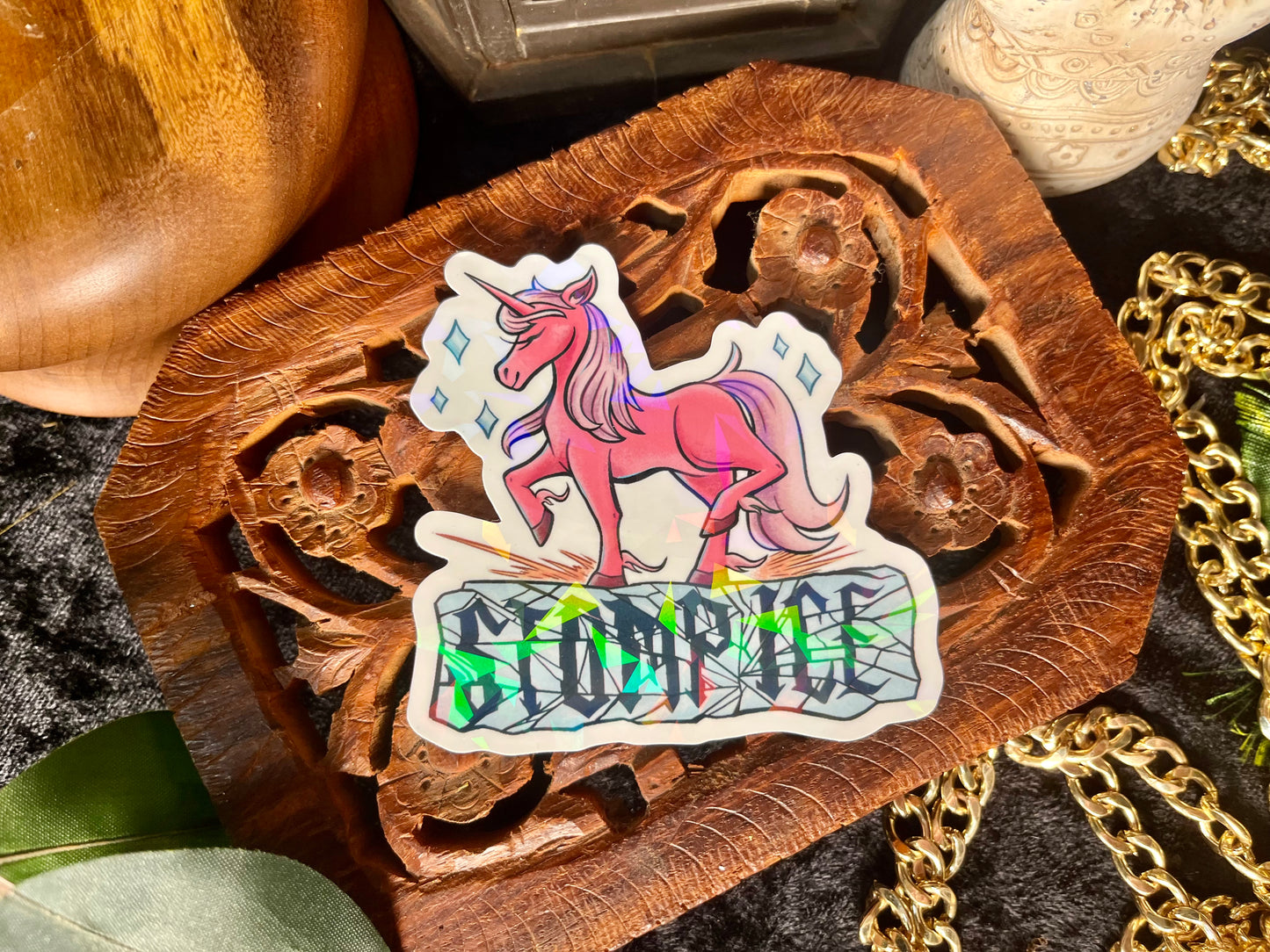 Holographic sticker with a unicorn design saying "Stomp ICE" on a wooden surface with gold chains and eclectic props