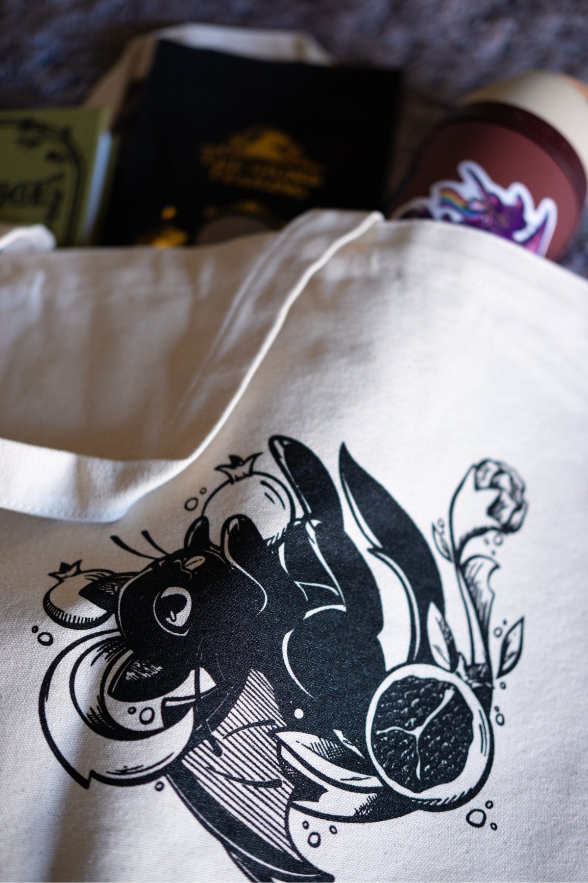 White tote bag with black design of a bat cat holding a pomegranate on a textured surface with books and tea spilling out