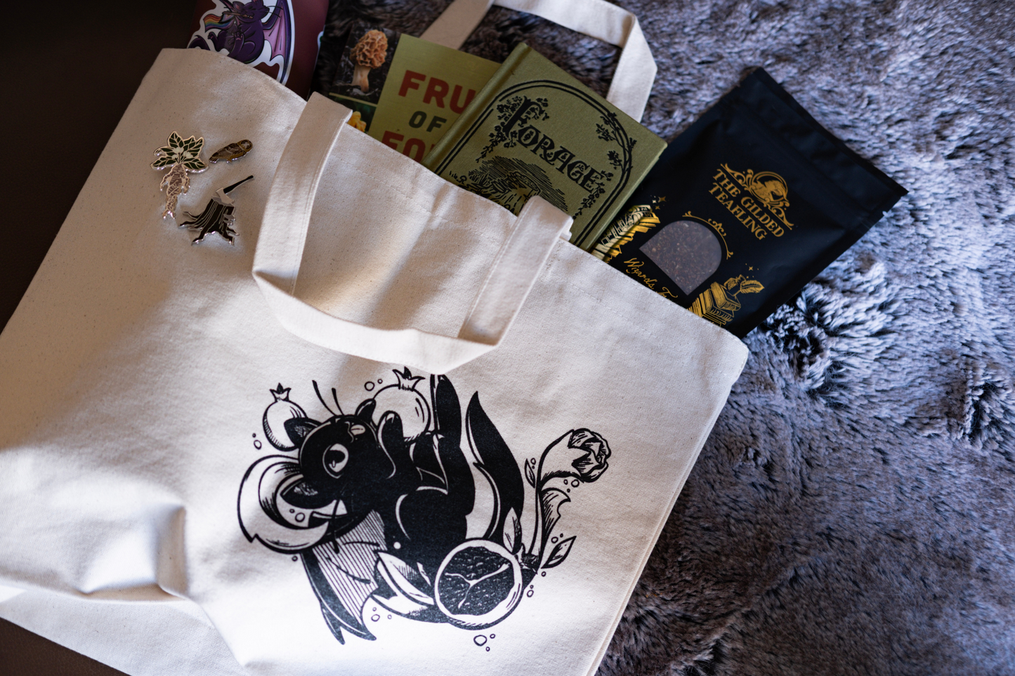 White tote bag with black design of a bat cat holding a pomegranate on a textured surface with books and tea spilling out