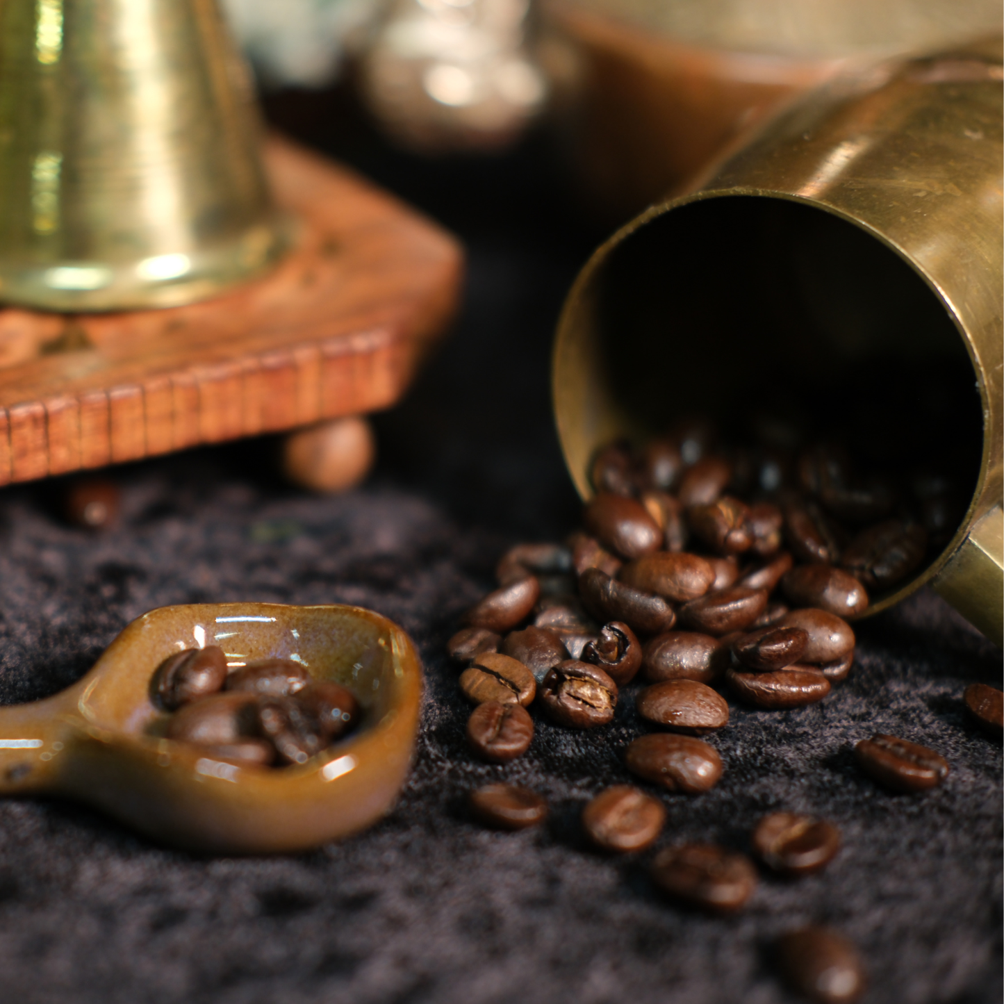 Coffee beans spilling from a brass scoop onto a dark surface with a wooden container in the background.