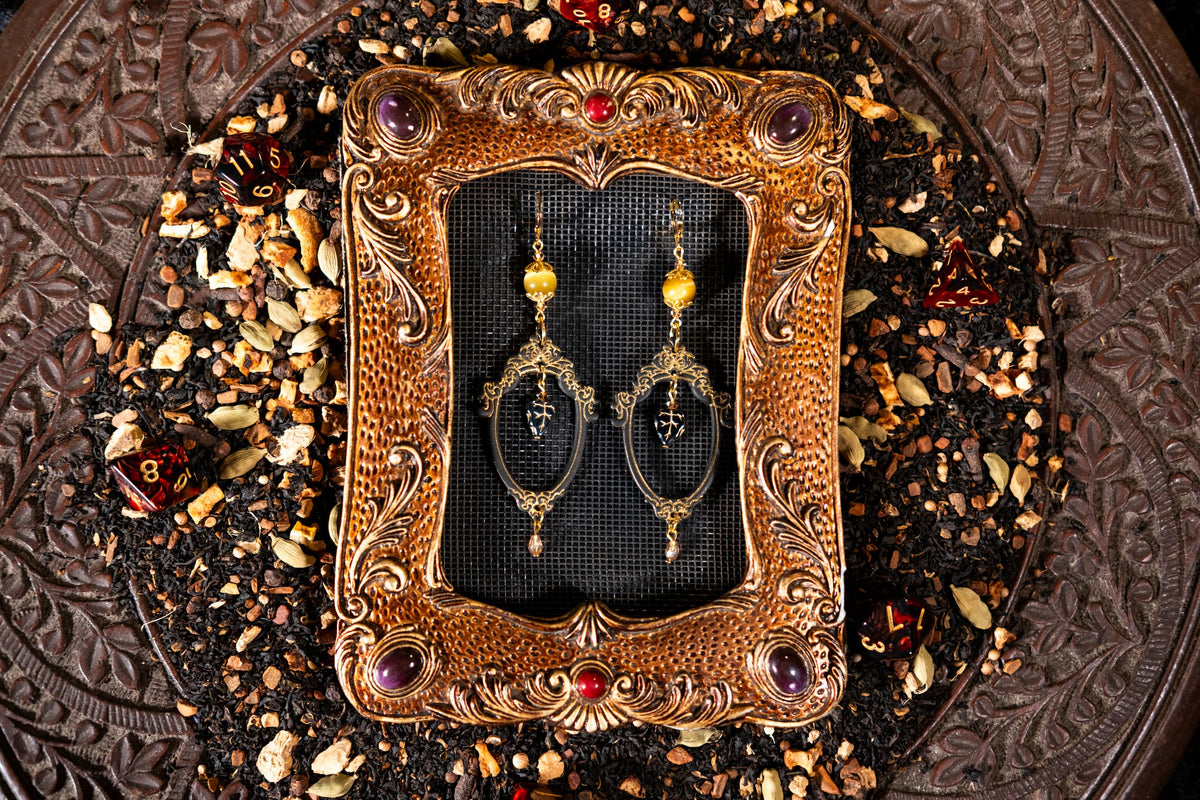 Ornate gold frame with black background displaying a pair of earrings on a textured surface.