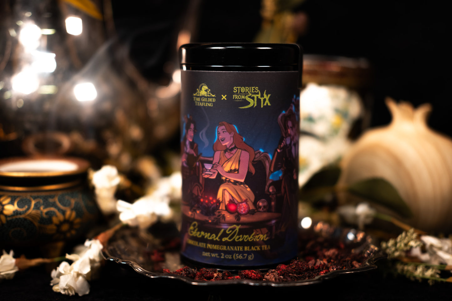 Black cylindrical tea tin with illustration depicting Hades and Persephone drinking tea with The Gilded Teafling and their bat cat, Assam