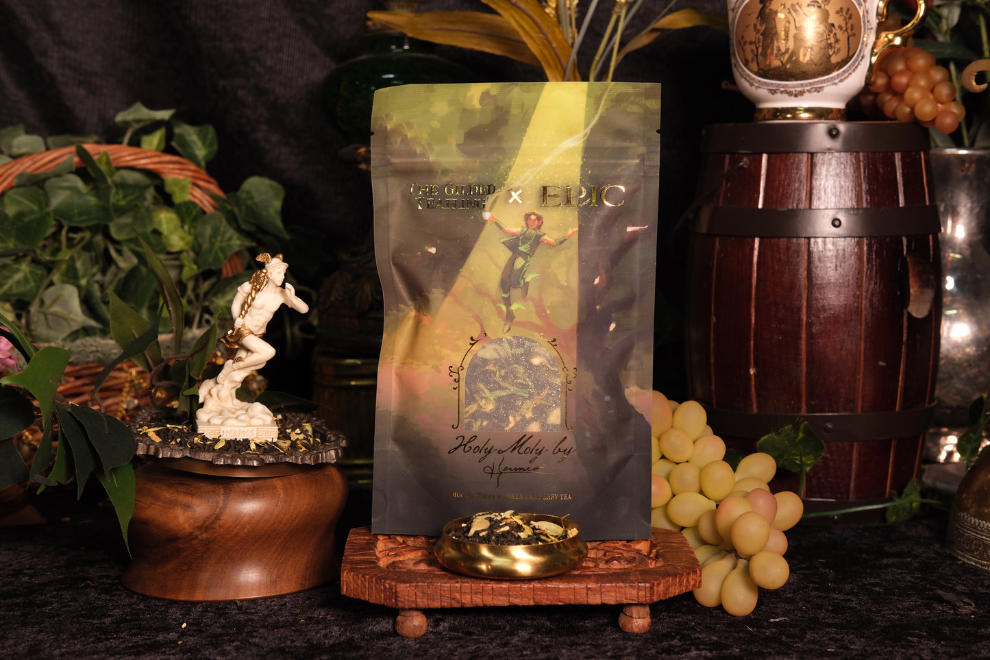 Tea packaging depicting Hermes floating down in a beam of light with a cup of tea in hand. Bag is set against a dark backdrop with Greek mythology related props surrounding it.