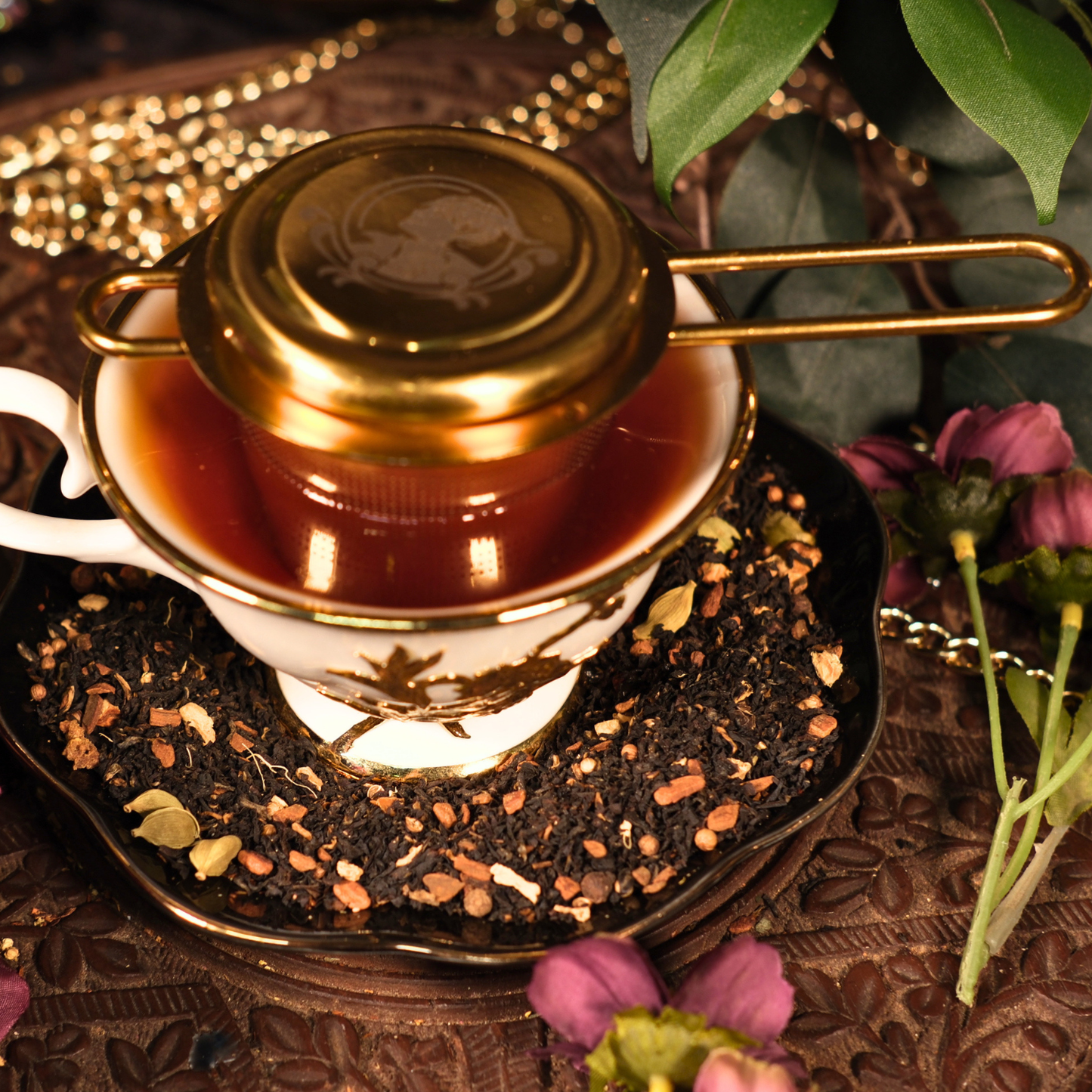 Gold tea strainer with lid sitting inside a white and gold teacup filled with amber-colored tea, sitting on a saucer filled with loose leaf chai