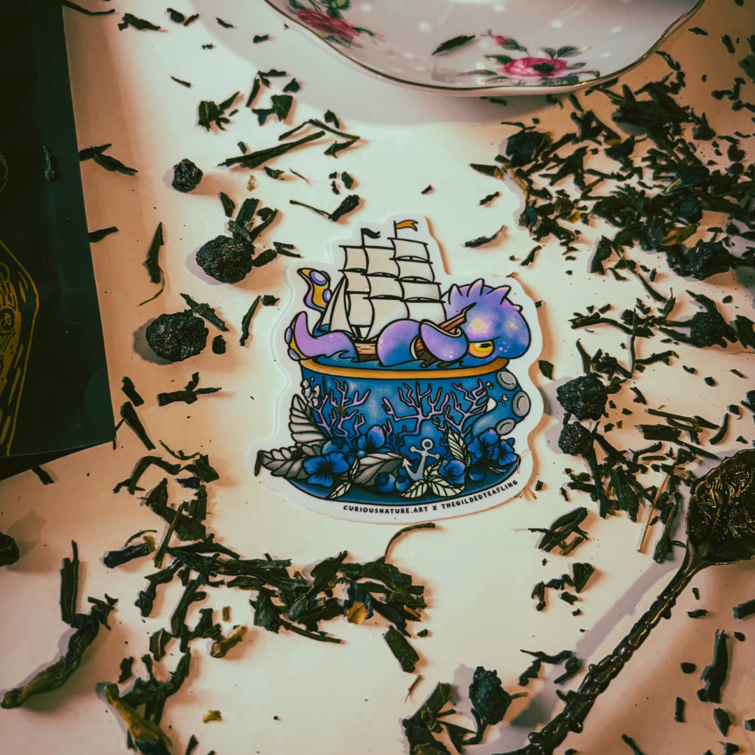 A die-cut sticker featuring an underwater scene with a purple octopus, blue fish, and a sailing ship on a pot, placed on a light surface surrounded by loose tea leaves.