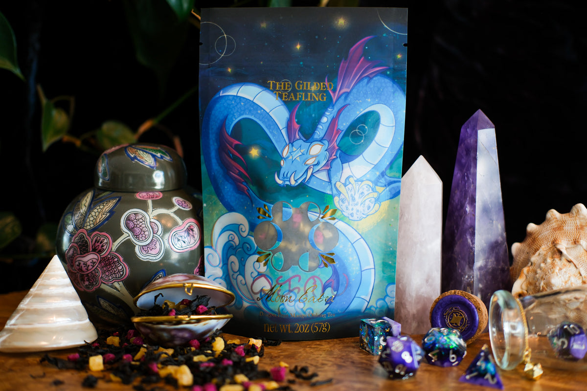 A teabag with a serpent dragon illustration stands on a wooden surface surrounded by crystals, a ceramic jar, seashells, loose tea pieces, and colorful dice in a dark styled setting.