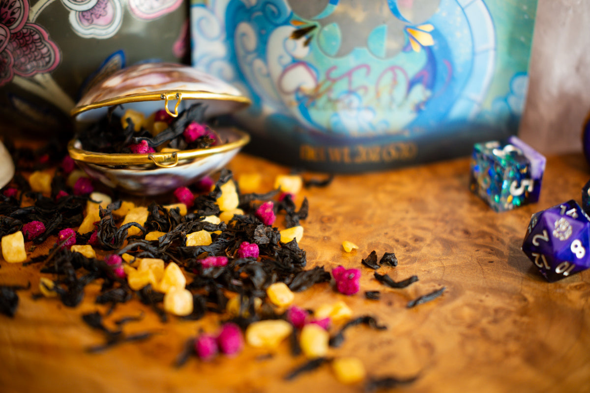 A styled tabletop scene with loose black tea leaves, yellow mango pieces, and pink freeze dried dragon fruit pieces scattered on a wooden surface. A small open shell-shaped tin filled with the tea mix sits in the foreground, with colorful polyhedral dice and a decorative blue illustrated container in the background.