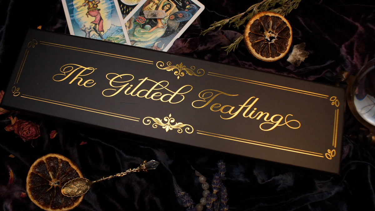 A black rectangular box with ornate gold script reading 'The Gilded Teaflings' lies on dark draped fabric, surrounded by dried orange slices, a spoon, tarot cards, and dried flowers.