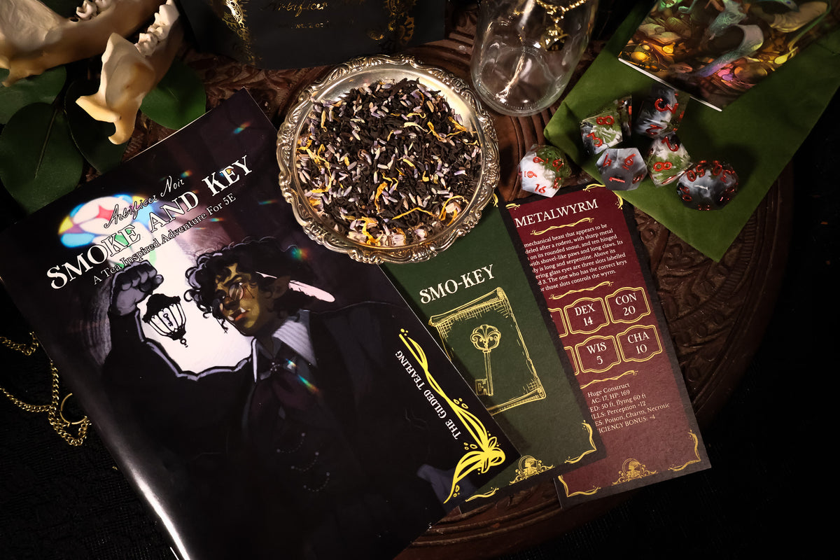 A styled tabletop flat-lay of a smoking card deck set, featuring a black 'Smoke and the Key' booklet, a green 'Smo-Key' card, a red 'Metalivyr' card, a silver dish of dried flower powder, and colorful polyhedral gaming dice on a dark wooden surface.