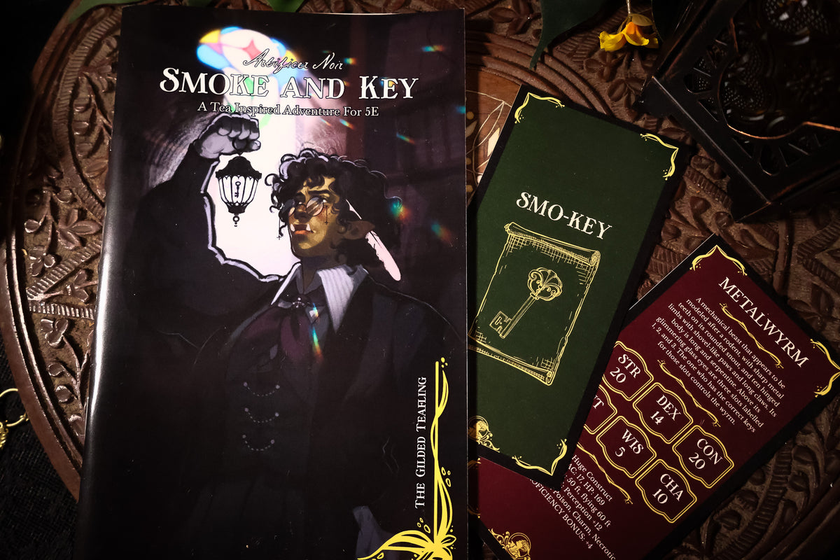 A tabletop flat-lay of a tabletop RPG supplement set titled 'Smoke and Key'. The set includes a black hardcover book with gothic artwork, a dark green card deck box labeled 'Smo-Key', and a dark red booklet labeled 'Metalwyrm'.