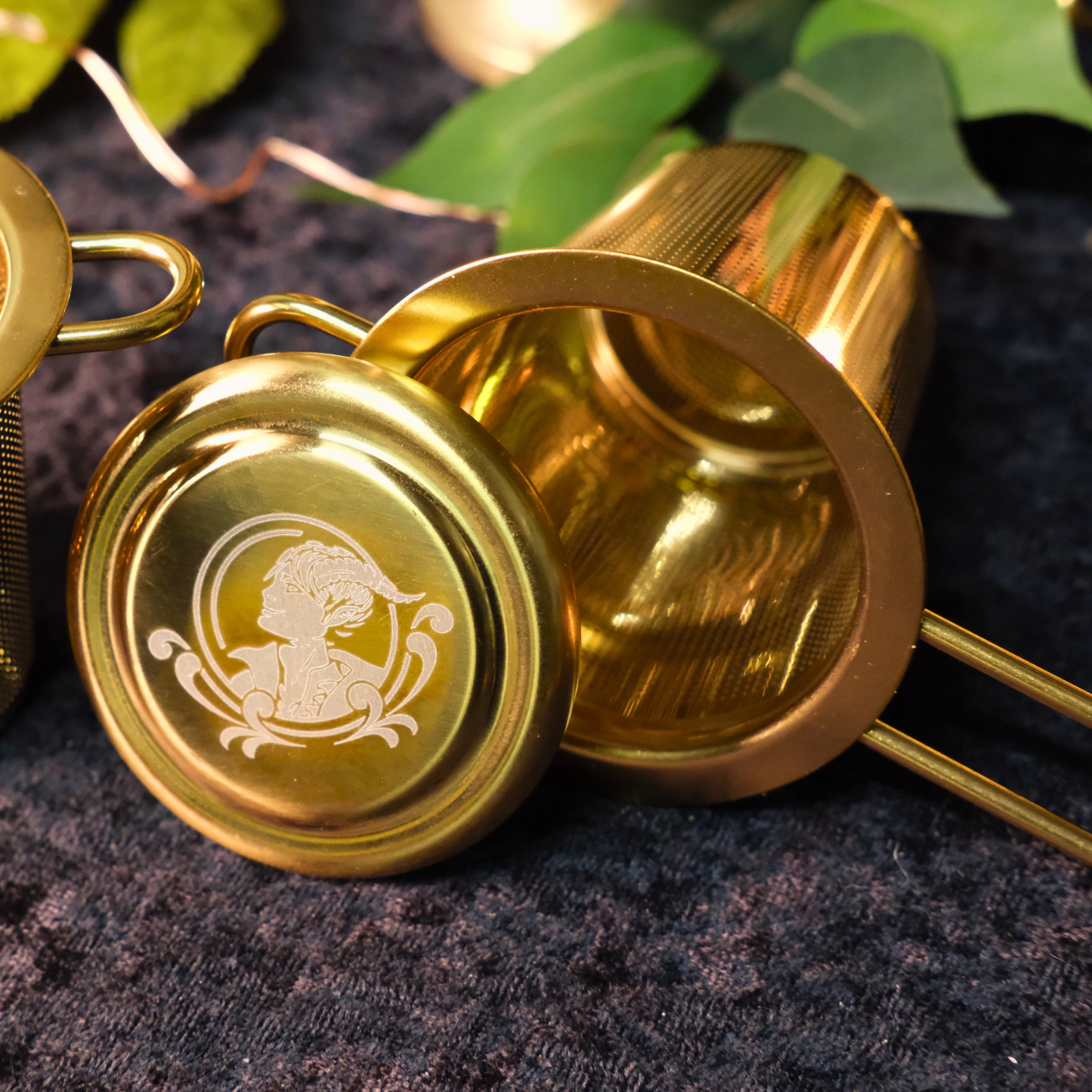 Gold metal strainer with engraved Tiefling head design on a dark textured surface with green leaves.